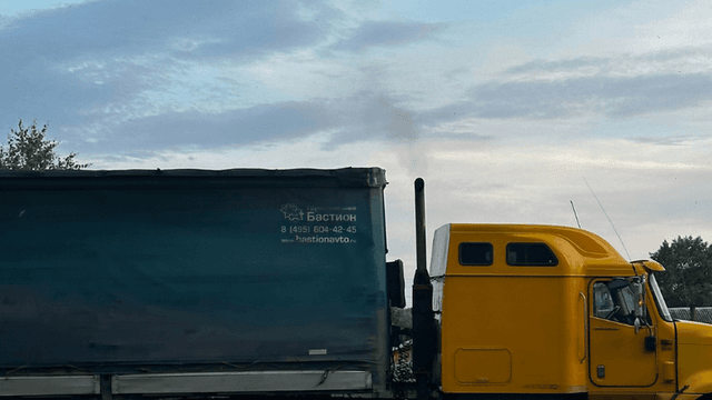 This image shows a yellow truck with a large, blue tarped trailer, with visible smoke coming from its exhaust pipe. The truck is marked with the text "Бастион" and includes a contact number and website, suggesting it's related to a transportation or logistics company. The truck is positioned in a rural or industrial setting, with a clear sky and some greenery in the background. The view focuses on the side of the truck, with the exhaust pipe releasing smoke into the air.