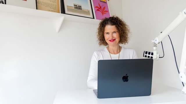 Dr Kate Armstrong seated behind a laptop with a podcast microphone in a bright home office.