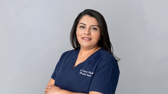 A woman in a blue medical uniform stands confidently against a gray background, smiling gently with arms crossed, conveying professionalism and warmth.