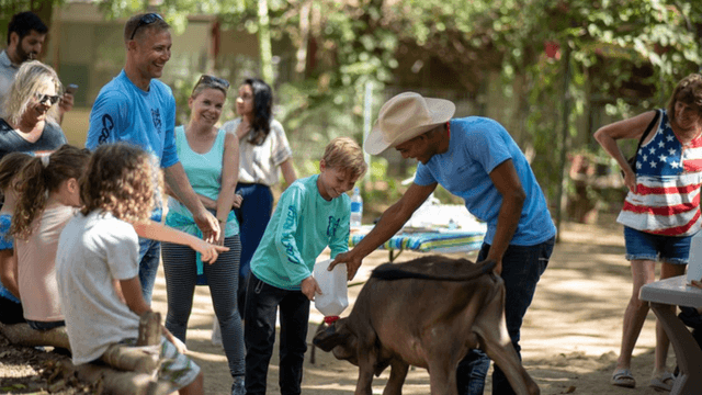 Children and adults smile while feeding a calf at The Monkey Farm’s community-focused recovery and healing center in Costa Rica.