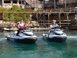 Two people ride Sea-Doo jet skis together on the water in front of Popeye Village.
