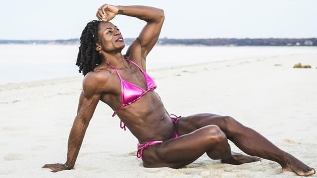 A strong, confident woman with muscular physique sits on a sandy beach, reflecting strength, discipline, and personal transformation.