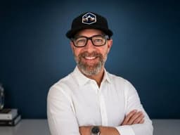 Blair Diamond wearing glasses and a black cap against a dark blue background.