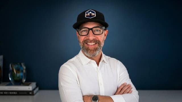 Blair Diamond wearing glasses and a black cap against a dark blue background.