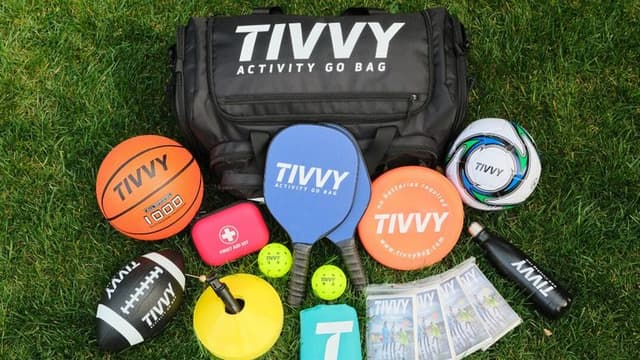 Black sports bag labeled "Tivvy" sits on grass surrounded by various activities gear: basketball, soccer ball, paddles, cones, water bottle, and discs.