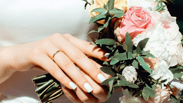 A close-up of a bride's hand holding a bouquet of pink and white flowers, showcasing her engagement ring and elegant manicure.