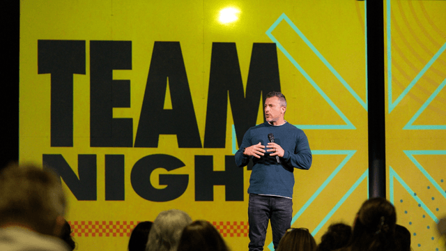Speaker presenting at a Team Night event, with a vibrant yellow backdrop and bold 'TEAM NIGHT' text. The speaker is engaging with the audience, emphasizing teamwork and motivation. The image captures a professional and energetic atmosphere at a leadership or team-building gathering.