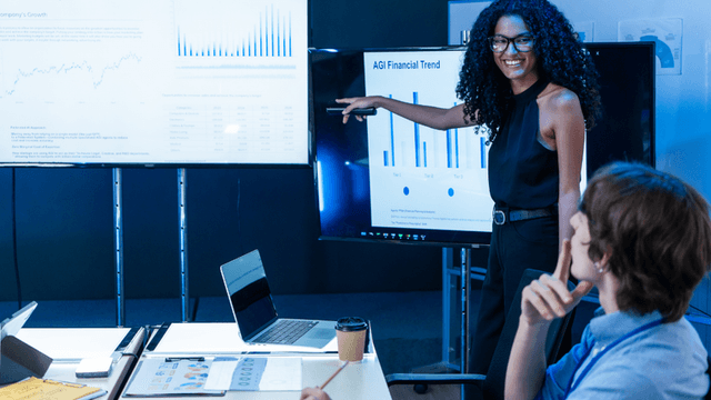 Presenter pointing to financial charts on digital displays during an AI business presentation.