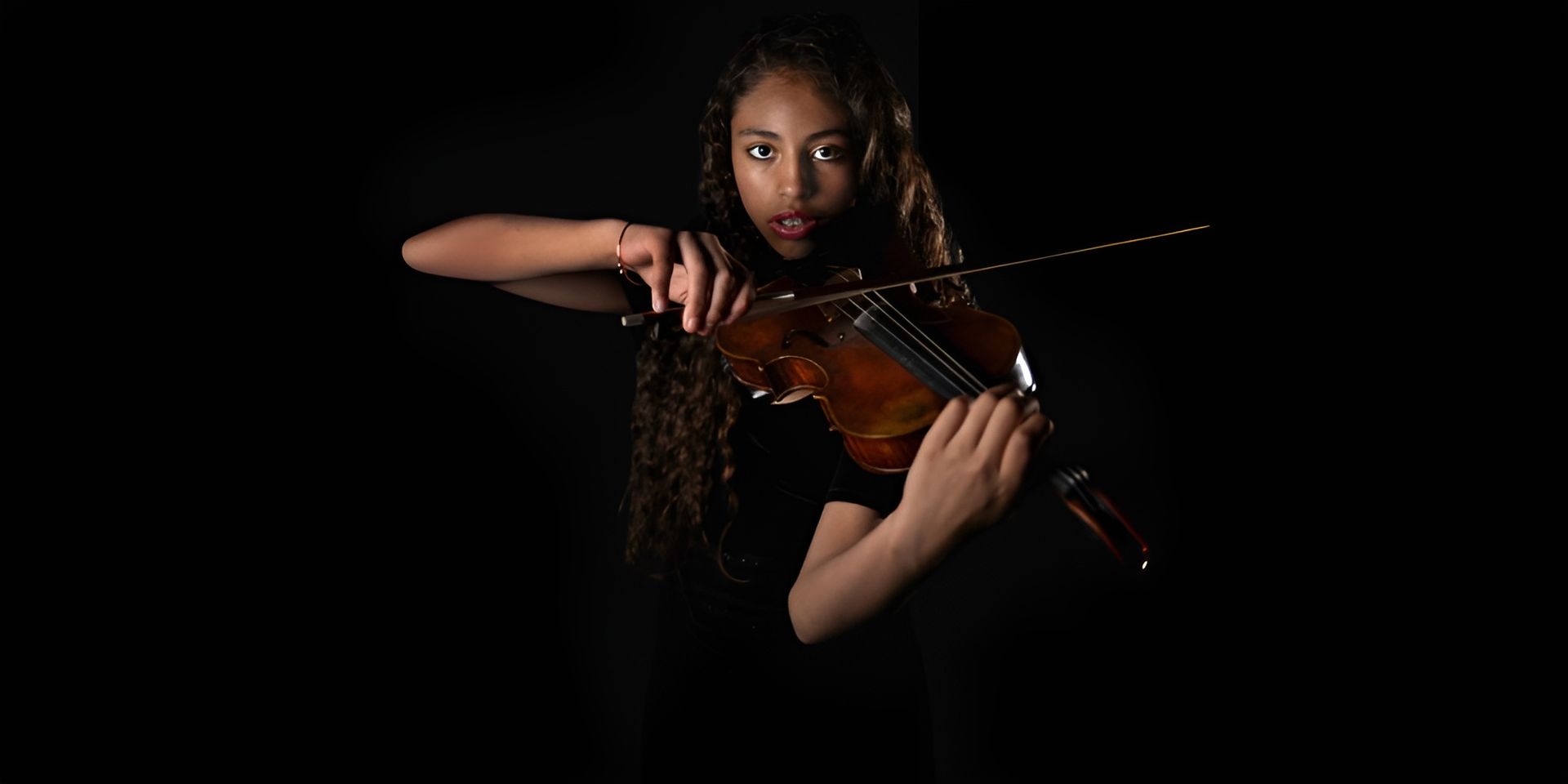 A young woman with curly hair, wearing a black top, playing the violin with intense focus against a dark background. A young woman with curly hair, wearing a black top, playing the violin with intense focus against a dark background.