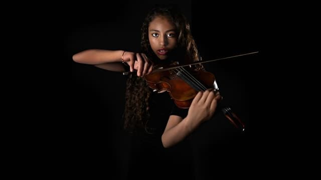 A young woman with curly hair, wearing a black top, playing the violin with intense focus against a dark background. A young woman with curly hair, wearing a black top, playing the violin with intense focus against a dark background.