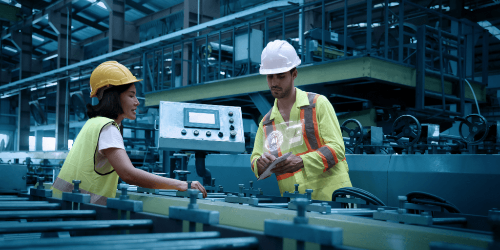 Two workers in a factory discussing plans near machinery and control panels