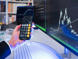 A person using a calculator app on a smartphone near a computer displaying graphs.