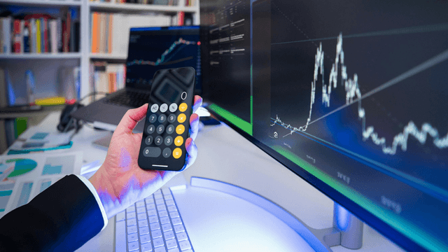 A person using a calculator app on a smartphone near a computer displaying graphs.