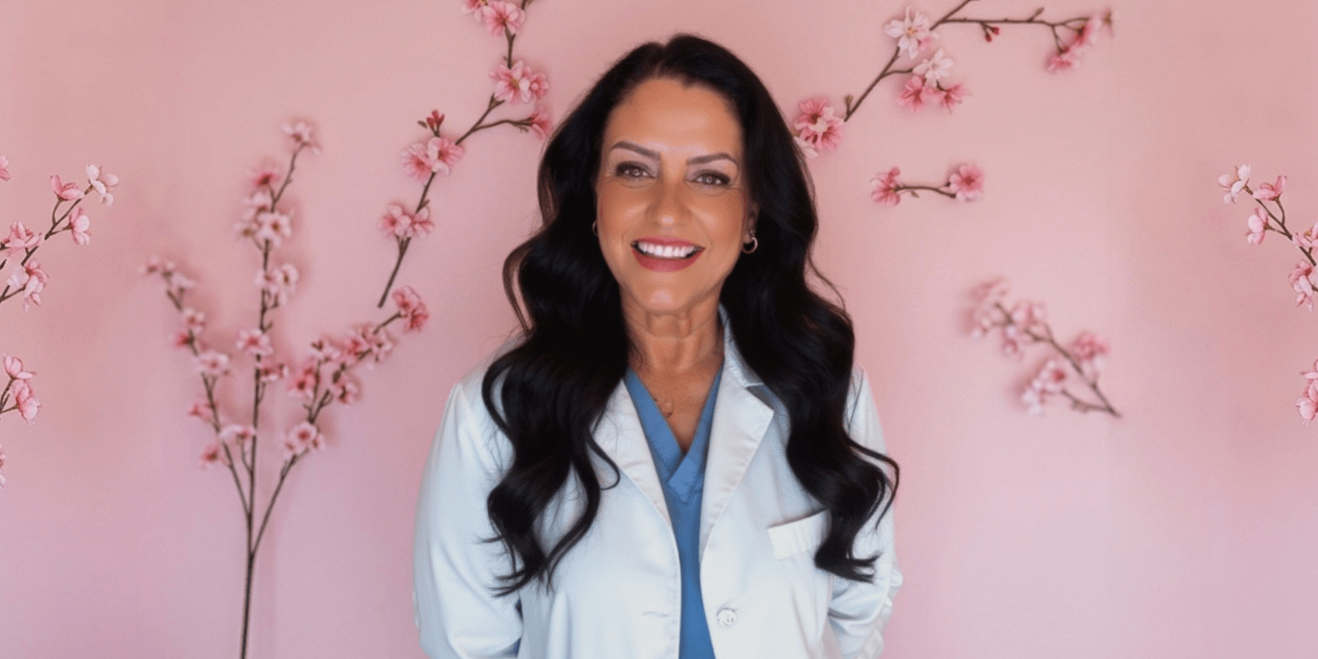 Smiling woman in a white coat stands against a pink floral background
