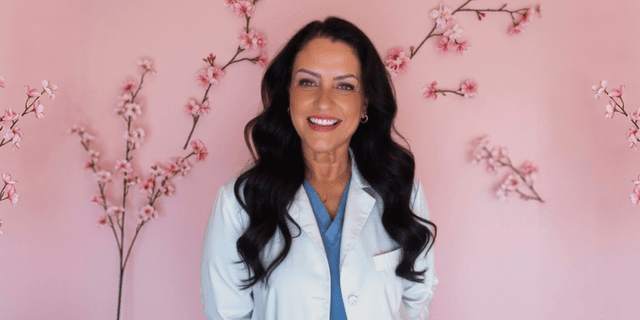 Smiling woman in a white coat stands against a pink floral background