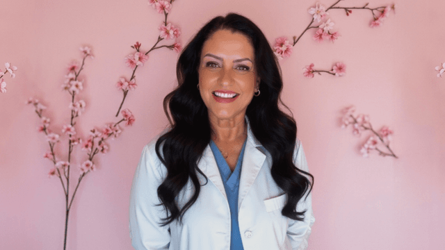 Smiling woman in a white coat stands against a pink floral background