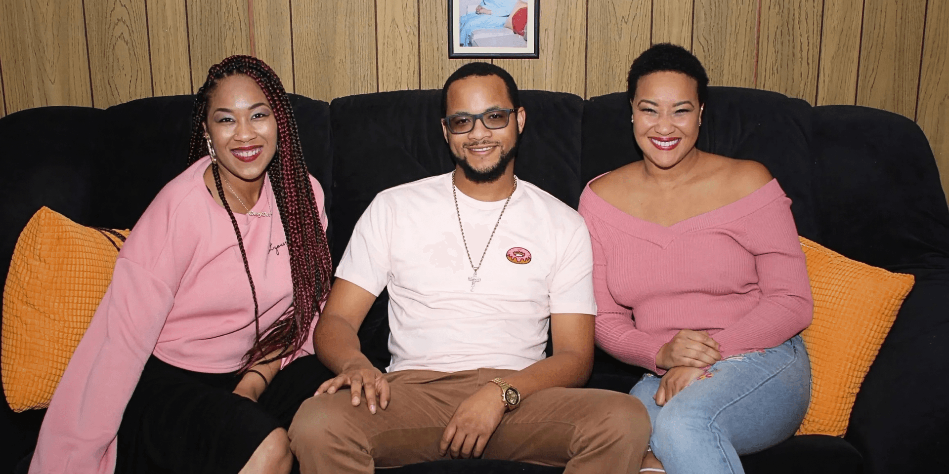 Three people sitting on a couch wearing pink clothing, smiling at the camera.