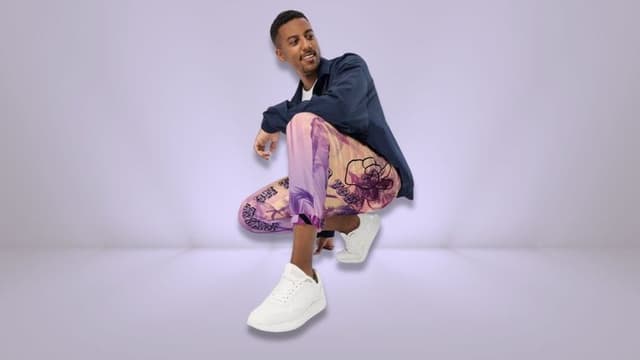 Man crouching in colorful patterned pants and white sneakers against a light purple background.