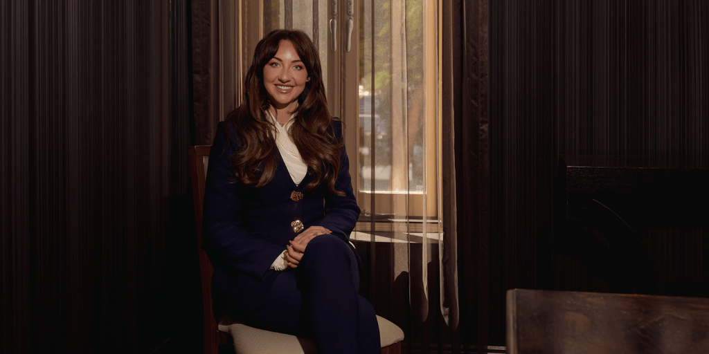 "Smiling woman in a blue suit sitting near a window, exuding confidence and warmth in a professional setting."