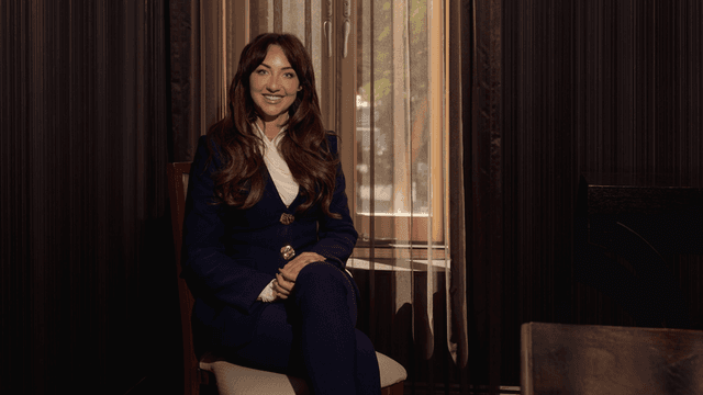"Smiling woman in a blue suit sitting near a window, exuding confidence and warmth in a professional setting."