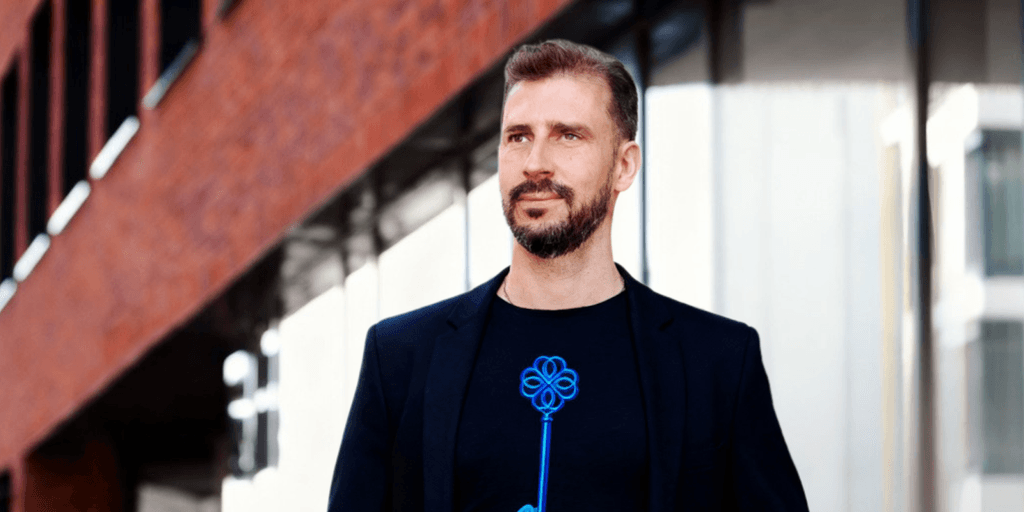 Peter Shogun Trnka in a black blazer and t-shirt with a blue key design, walking in an urban setting.