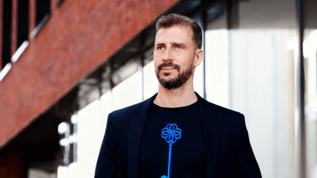 Peter Shogun Trnka in a black blazer and t-shirt with a blue key design, walking in an urban setting.
