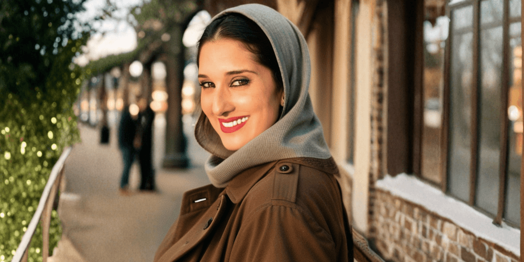 Vjolca Capri smiles confidently in a brown trench coat and gray scarf, with a blurred outdoor backdrop and warm lighting.