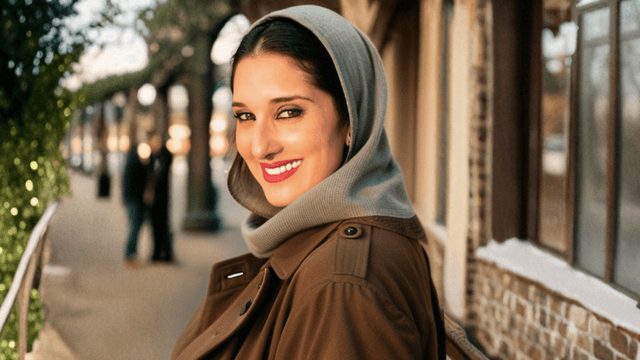 Vjolca Capri smiles confidently in a brown trench coat and gray scarf, with a blurred outdoor backdrop and warm lighting.