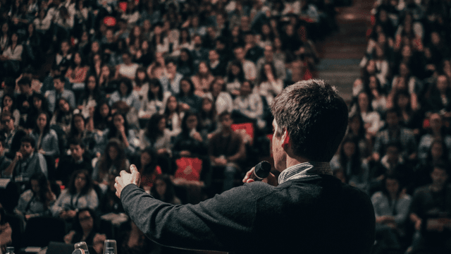 The image shows a speaker addressing a large audience at a conference or seminar, highlighting the importance of communication.