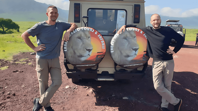 The image shows two individuals standing beside a safari vehicle with "Ernest Magic Tours & Safaris" logo, set in Tanzania.