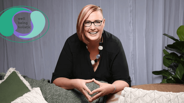 "Smiling woman in black shirt and colorful necklace, laughing while sitting on a couch surrounded by pillows, with the 'Well Being Holistic' logo in the corner."