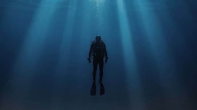 A diver immersed in the calm, deep blue of the ocean, illuminated by rays of sunlight filtering through the water.