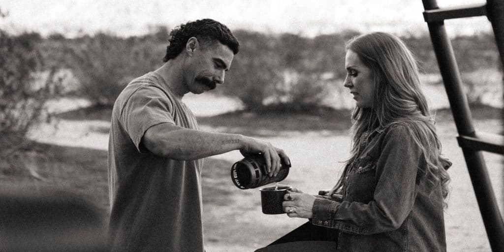 A man pours coffee into a mug held by a woman outdoors. The scene conveys a peaceful and warm moment, set against a backdrop of blurred natural scenery.
