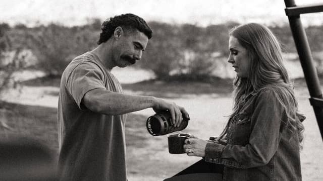 A man pours coffee into a mug held by a woman outdoors. The scene conveys a peaceful and warm moment, set against a backdrop of blurred natural scenery.
