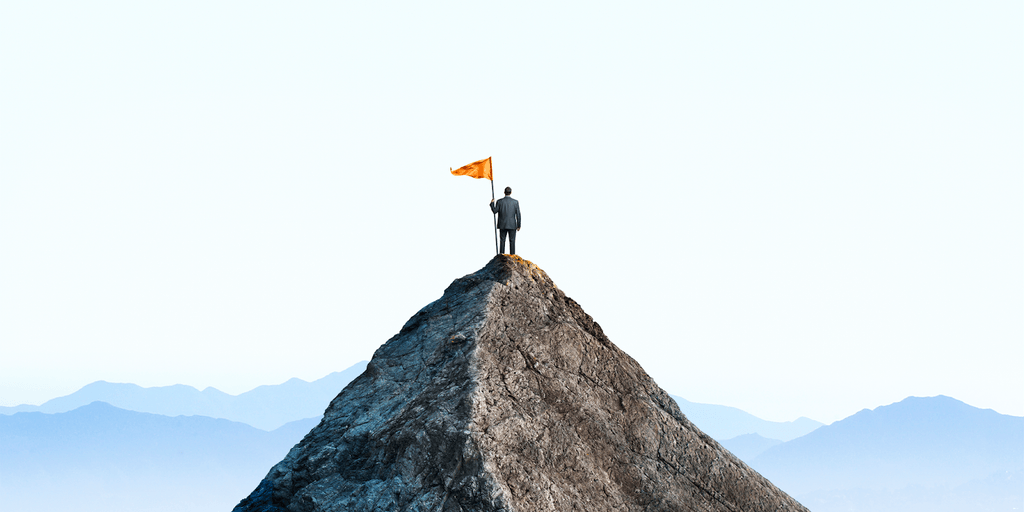 A person in a suit stands on a mountain peak, holding an orange flag, symbolizing personal transformation and success