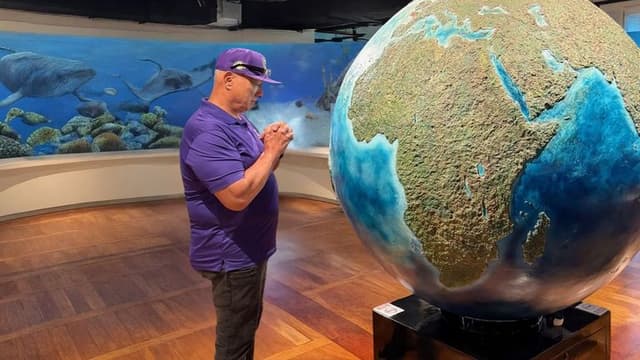 Apostle Constantine I. Nightingdale stands by a large globe, contemplating with hands clasped in a museum, surrounded by an underwater mural featuring humpback whales.