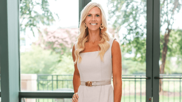 Professional woman smiling in a light-colored dress standing near large windows with greenery outside.