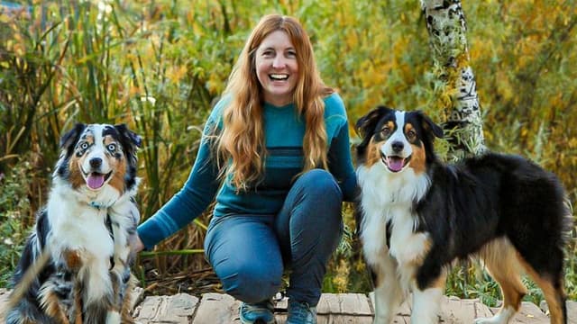 A woman smiles with two Australian Shepherd dogs in a natural setting, reflecting the bond built through expert training.