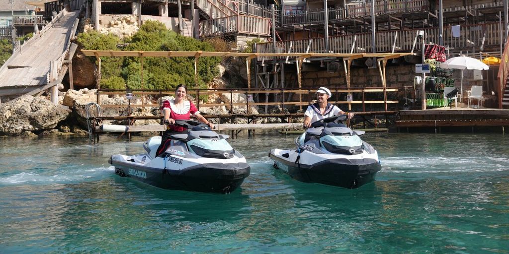 Two people ride Sea-Doo jet skis together on the water in front of Popeye Village.