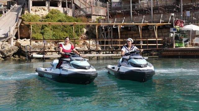 Two people ride Sea-Doo jet skis together on the water in front of Popeye Village.