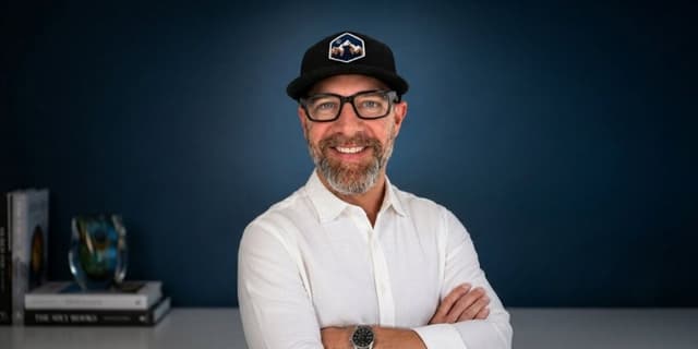 Blair Diamond wearing glasses and a black cap against a dark blue background.