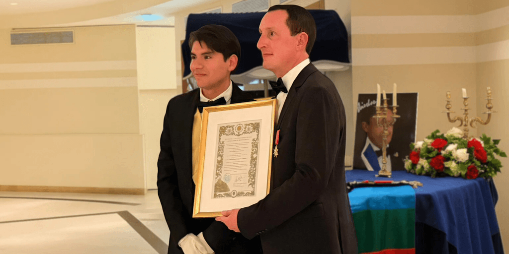 "Prince Antoine V (Laurent LAFAYNE) receiving an award with a Mapuche flag in the background."