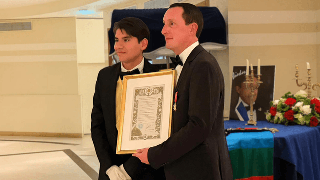 "Prince Antoine V (Laurent LAFAYNE) receiving an award with a Mapuche flag in the background."