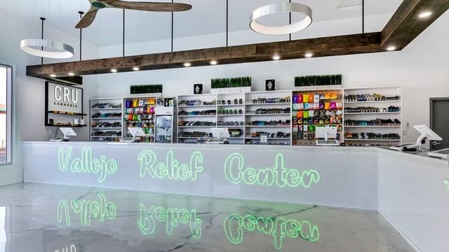 Spacious, modern dispensary interior with sleek white counters and shelves stocked with colorful products. Neon sign reads "Vallejo Relief Center."