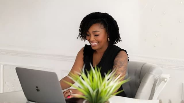 A smiling woman with curly hair works on a laptop, surrounded by plants, possibly related to entrepreneurship or healthcare.