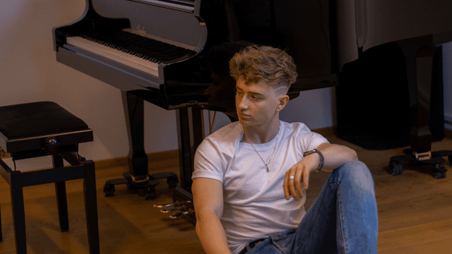 "Young male musician sitting on the floor next to a grand piano, wearing a white t-shirt and jeans, looking contemplative with his hand resting on his knee, exuding a relaxed and thoughtful atmosphere."