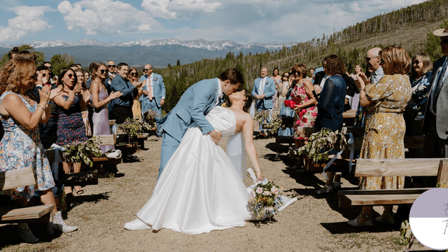 A wedding couple sharing a kiss during their outdoor ceremony, surrounded by guests cheering, with a stunning mountain backdrop.