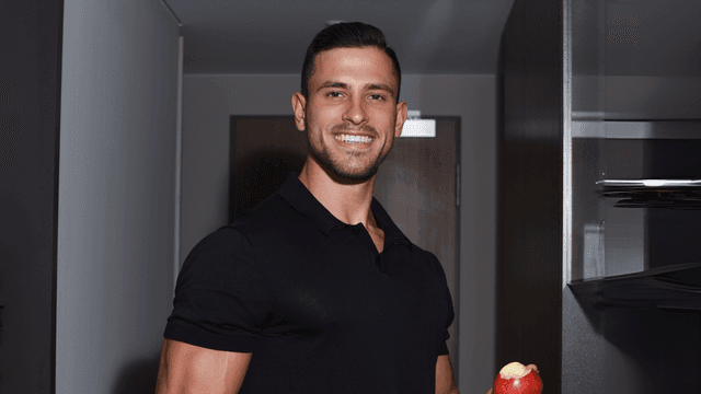"Man in a black polo shirt holding a red apple, smiling in a modern kitchen setting with shelves in the background."