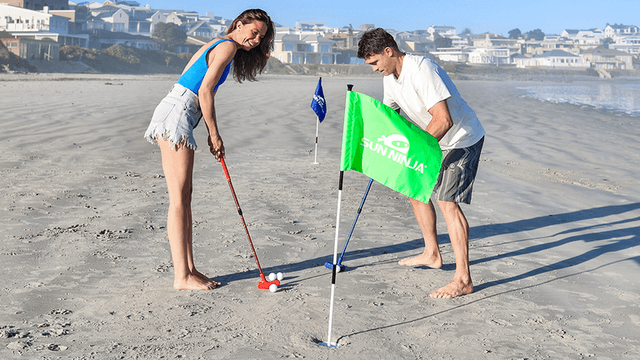 "Couple enjoying beach golf with colorful flags and golf clubs, one of them playing with a red club and the other adjusting the green flag."