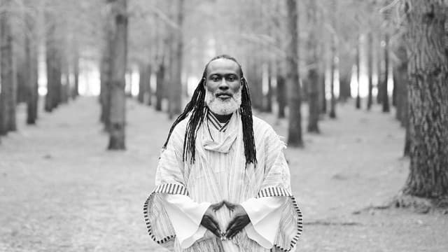 Black-and-white photo of SPEARHEART standing in a forest, wearing traditional patterned robe, looking directly at the camera.